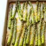 Garlic parmesan roasted asparagus - the image shows a baking tray with cooked asparagus spears on it. the tray is rectangular in shape and is placed on a white countertop. the spears are arranged in a single layer on the tray, with some of them overlapping each other. they are covered in a golden-brown sauce, which appears to be seasoned with herbs and spices. the sauce is drizzled over the spears, giving them a glossy sheen. the overall appearance of the dish is fresh and appetizing.