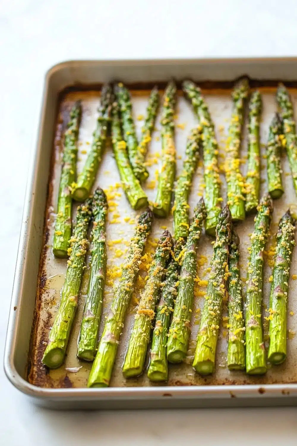 Garlic parmesan roasted asparagus - the image shows a baking tray with cooked asparagus spears on it. the tray is rectangular in shape and is placed on a white countertop. the spears are arranged in a single layer on the tray, with some of them overlapping each other. they are covered in a golden-brown sauce, which appears to be seasoned with herbs and spices. the sauce is drizzled over the spears, giving them a glossy sheen. the overall appearance of the dish is fresh and appetizing.