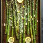 Garlic parmesan roasted asparagus - the image shows a baking tray filled with cooked asparagus spears. the spears are arranged in a single layer on the tray, with some of them overlapping each other. the tray is lined with parchment paper and there are several lemon wedges scattered around the tray. the background is a dark grey countertop. the overall appearance of the dish is fresh and appetizing.