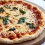 Gluten free pizza crust caputo - the image is of a freshly baked pizza on a wooden cutting board. the pizza has a golden brown crust and is topped with melted cheese and fresh basil leaves. the cheese is golden brown and appears to be melted and bubbly. the basil leaves are scattered across the top of the pizza, adding a pop of green color to the dish. the cutting board is placed on a white marble countertop.