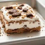 Graham bar cookie and cream - the image shows a slice of ice cream cake on a white rectangular baking tray. the cake has a layer of white frosting on top, with a crumbly texture on the sides. the frosting is drizzled over the top of the cake, and there are small chocolate chips scattered around the edges. the tray is sitting on a wooden table, and the background is blurred.