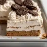 Graham bar cookie and cream - the image shows a slice of oreo cheesecake on a white rectangular baking tray. the cheesecake has a golden brown crust and is topped with a generous layer of whipped cream. there are several oreo cookies scattered on top of the cheesecake, adding a pop of color to the dish. the tray is sitting on a marble countertop.