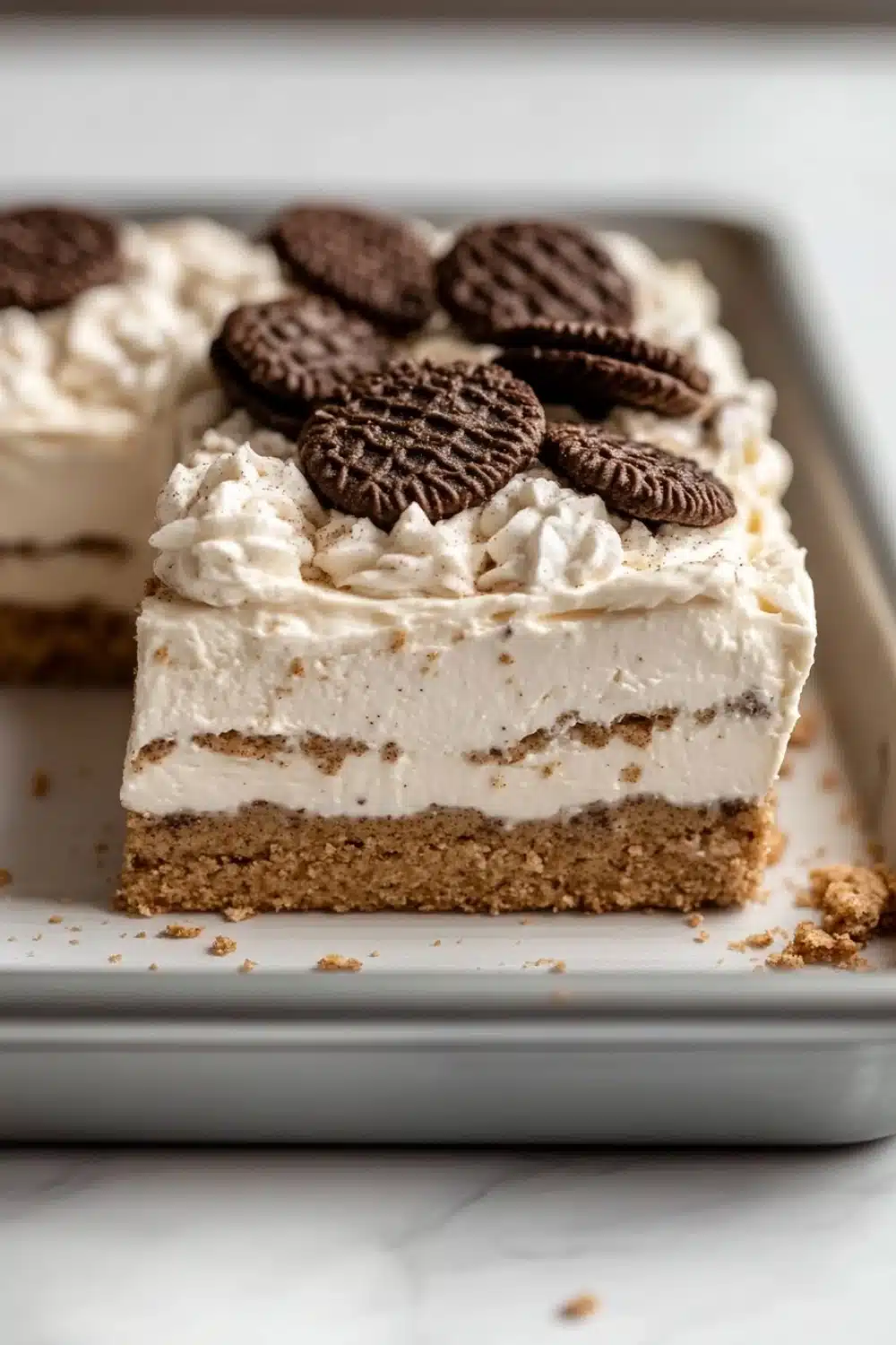 Graham bar cookie and cream - the image shows a slice of oreo cheesecake on a white rectangular baking tray. the cheesecake has a golden brown crust and is topped with a generous layer of whipped cream. there are several oreo cookies scattered on top of the cheesecake, adding a pop of color to the dish. the tray is sitting on a marble countertop.