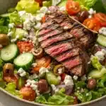 Greek salad with lamb - the image is a close-up of a large bowl of salad. the salad is made up of various types of vegetables, including lettuce, cherry tomatoes, cucumbers, olives, and feta cheese. in the center of the salad, there is a large piece of grilled steak that appears to be seasoned with herbs and spices. the steak is cut into thin slices and is placed on top of the lettuce. the bowl is white and has a textured surface. the background is blurred, so the focus is on the salad.