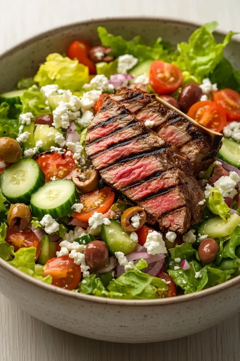 Greek salad with lamb - the image is a close-up of a large bowl of salad. the salad is made up of various types of vegetables, including lettuce, cherry tomatoes, cucumbers, olives, and feta cheese. in the center of the salad, there is a large piece of grilled steak that appears to be seasoned with herbs and spices. the steak is cut into thin slices and is placed on top of the lettuce. the bowl is white and has a textured surface. the background is blurred, so the focus is on the salad.