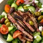 Greek salad with lamb - the image is a close-up of a salad in a bowl. the salad is made up of various types of vegetables, including lettuce, cherry tomatoes, cucumbers, olives, red onions, and feta cheese. in the center of the salad, there are slices of grilled steak arranged in a circular pattern. the steak appears to be seasoned with herbs and spices, and the vegetables are cooked to perfection. the bowl is made of light-colored ceramic and is sitting on a white marble countertop.