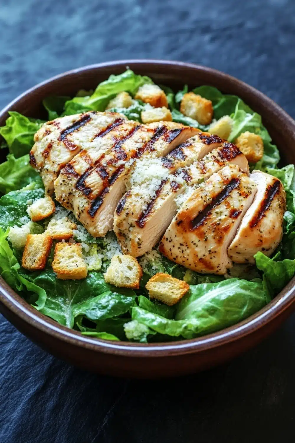 Grilled chicken caesar salad - the image is a close-up of a bowl of salad. the salad is made up of green lettuce leaves and is topped with grilled chicken breasts. the chicken breasts are golden brown and appear to be seasoned with herbs and spices. there are also croutons scattered throughout the salad, adding a crunchy texture. the bowl is made of dark brown ceramic and is sitting on a dark blue tablecloth.