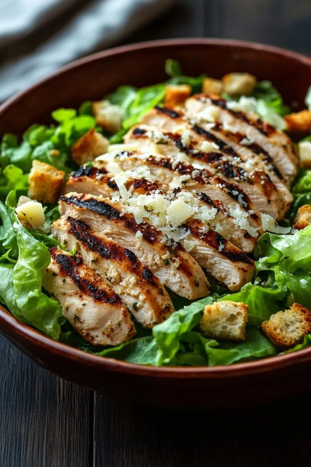 Grilled chicken caesar salad - the image is a close-up of a salad in a brown bowl. the salad is made up of lettuce leaves and croutons, and there are several pieces of grilled chicken arranged in a circular pattern on top. the chicken appears to be seasoned with herbs and spices, and is drizzled with grated parmesan cheese. the bowl is sitting on a wooden table, and the background is blurred, making the salad the focal point of the image.