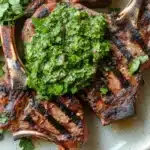 Grilled lamb chops with chimichurri - the image shows a plate of grilled steak with a generous amount of green salsa on top. the steak appears to be medium-rare and has a dark brown crust on the outside and a juicy center. the salsa is made up of chopped cilantro and herbs, and it is drizzled over the top of the steak. the plate is white with a light blue rim, and the steak is arranged in a circular pattern on the plate. the background is a wooden table.