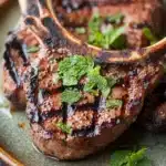 Grilled lamb chops with mint - the image shows a plate of grilled pork chops. the pork chops are cooked to a golden brown color and have a crispy texture. they are garnished with fresh mint leaves, adding a pop of green color to the dish. the plate is made of dark green ceramic and is placed on a wooden table. the background is blurred, making the pork chops the focal point of the image.