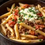Ground beef and pasta skillet - the image shows a bowl of pasta dish on a wooden table. the pasta appears to be penne, which is a type of penne pasta, and it is topped with ground beef, tomato sauce, and a dollop of sour cream. the dish is garnished with chopped parsley. the bowl is black and has a handle on one side. in the background, there is another bowl of french fries. the overall color scheme of the dish is warm and inviting.