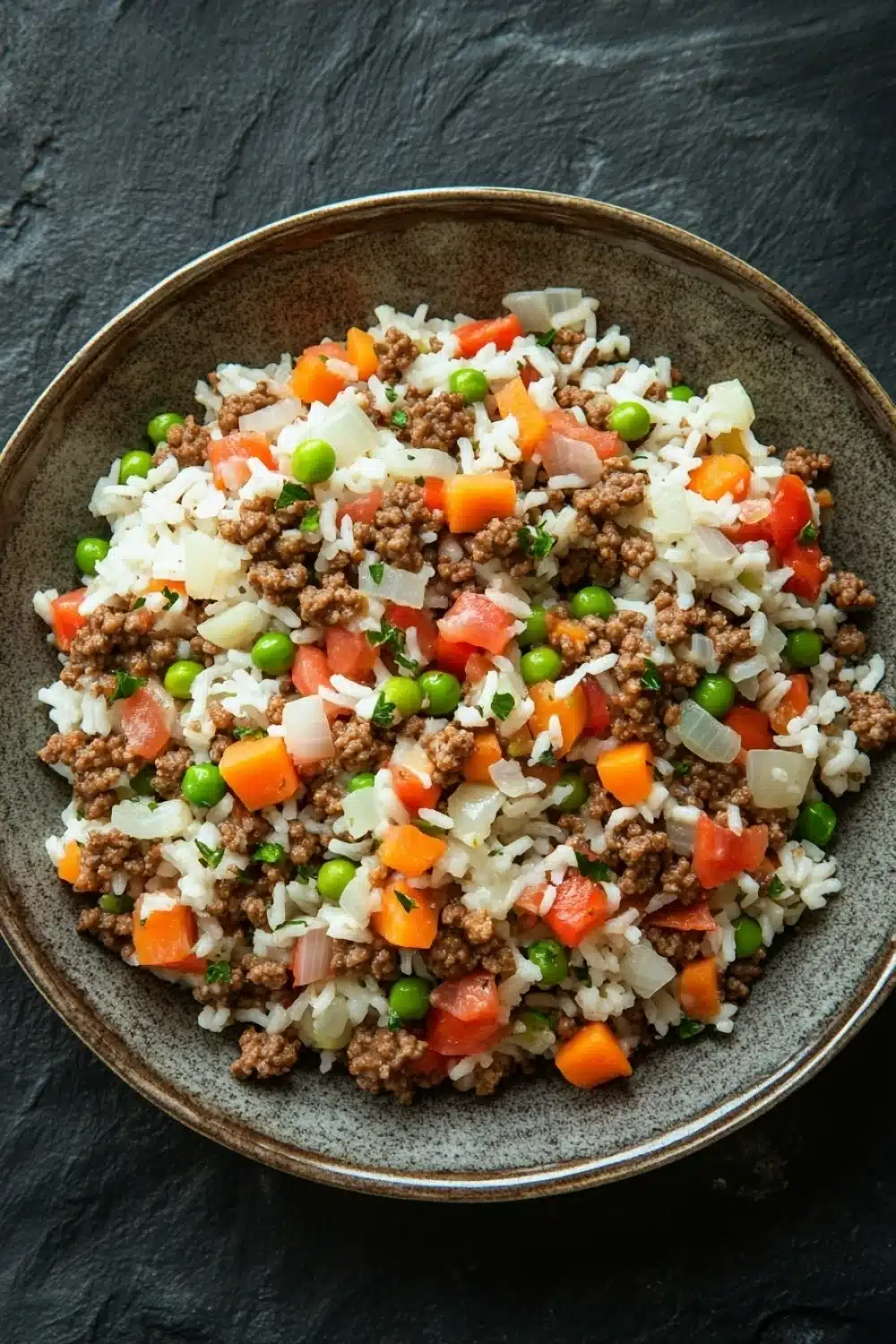 Ground beef and rice - the image is a close-up of a bowl of food. the bowl is made of dark brown ceramic and is placed on a dark grey textured surface. the food in the bowl appears to be a mix of rice, ground beef, diced carrots, peas, and onions. the rice is white and fluffy, and the ground beef is a light brown color. the vegetables are bright orange and green, and they are scattered throughout the dish. the dish looks fresh and appetizing.