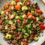 Ground beef and zucchini - the image is a close-up of a bowl of food. the bowl is made of light-colored ceramic with a speckled pattern. the food appears to be a salad with a variety of ingredients, including ground beef, zucchini, tomatoes, onions, and chopped herbs. the vegetables are arranged in a colorful and appetizing manner, with some overlapping each other. the dish is garnished with fresh parsley, adding a pop of green color to the dish. the background is a white wooden surface, and there are a few sprigs of parsley scattered around the bowl.