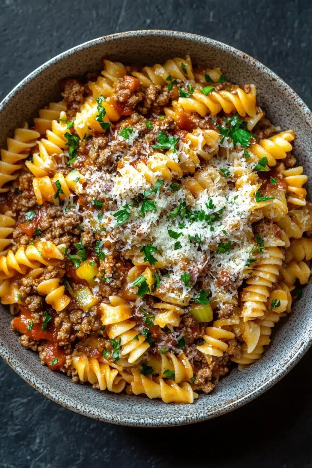 Ground beef with pasta - the image is a close-up of a bowl of pasta dish. the pasta appears to be fusilli, which is a type of spiral-shaped pasta. the dish is made with ground beef, tomato sauce, and chunks of meat. the meat is cooked in a tomato-based sauce and is garnished with chopped parsley. there is also a sprinkle of grated parmesan cheese on top of the dish, adding a pop of green color. the bowl is made of ceramic and is sitting on a dark grey countertop.