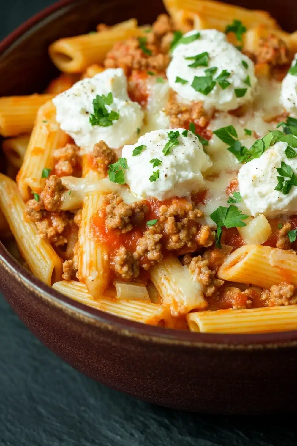 Ground turkey for dinner casseroles pasta - the image is a close-up of a bowl of pasta dish. the pasta appears to be penne, which is a type of penne pasta, and it is cooked in a tomato-based sauce. the dish is topped with chunks of ground meat, possibly ground beef, and a dollop of sour cream. the sauce is a deep red color and is drizzled over the top of the pasta. there are also a few sprigs of fresh parsley scattered throughout the dish, adding a pop of green color. the bowl is made of dark brown ceramic and is sitting on a dark blue surface.