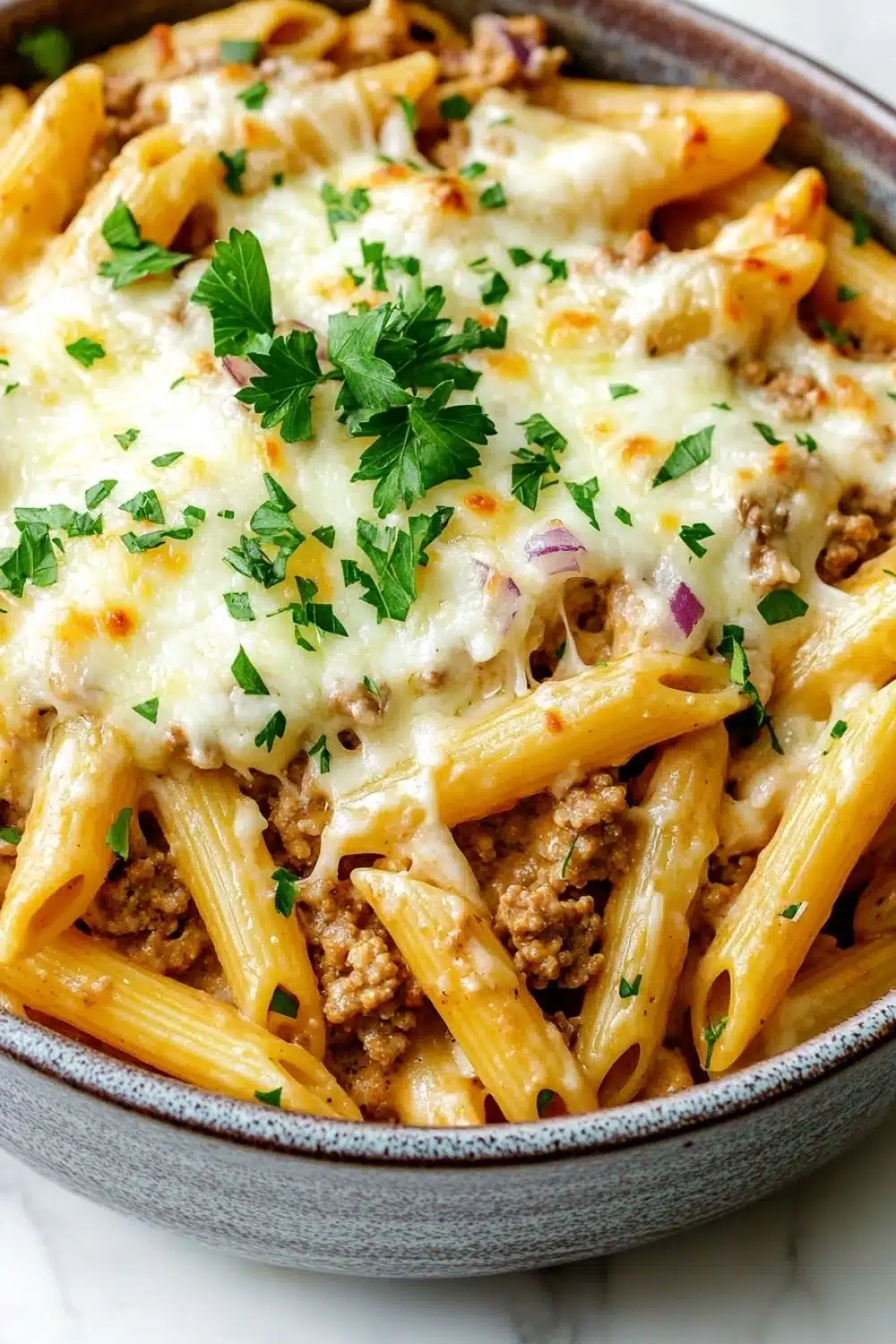 Ground turkey for dinner casseroles pasta - the image shows a bowl of penne pasta dish. the pasta is cooked al dente and is topped with a layer of ground beef and melted cheese. the dish is garnished with fresh parsley and red onions. the bowl is made of ceramic and is sitting on a white marble countertop.