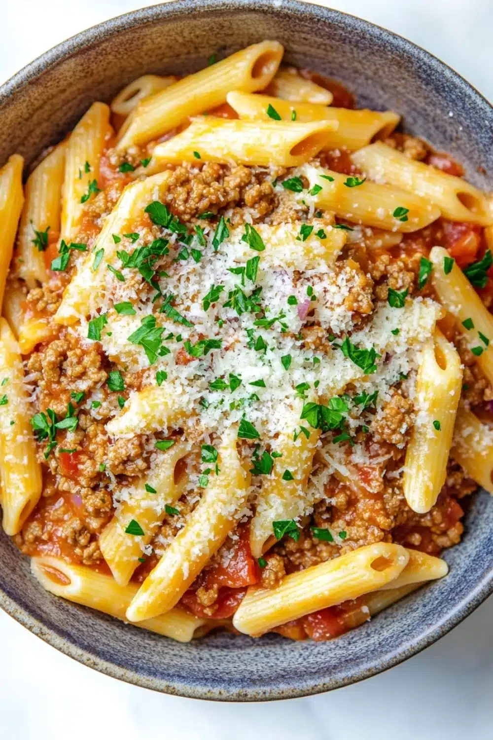 Ground turkey for dinner pasta - the image is a close-up of a bowl of pasta dish. the pasta appears to be penne, which is a type of penne pasta, and it is cooked in a tomato-based sauce with chunks of ground meat and tomato sauce. the dish is garnished with chopped parsley and grated parmesan cheese. the bowl is made of ceramic and has a dark blue rim. the background is white, making the colors of the dish stand out.