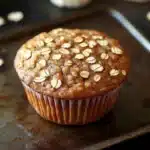 High protein banana oat muffin - the image shows a freshly baked muffin on a baking tray. the muffin is golden brown in color and has a crumbly texture. it is topped with chopped oats, giving it a crunchy appearance. in the background, there are several whole bananas scattered around the tray, adding to the rustic feel of the image.