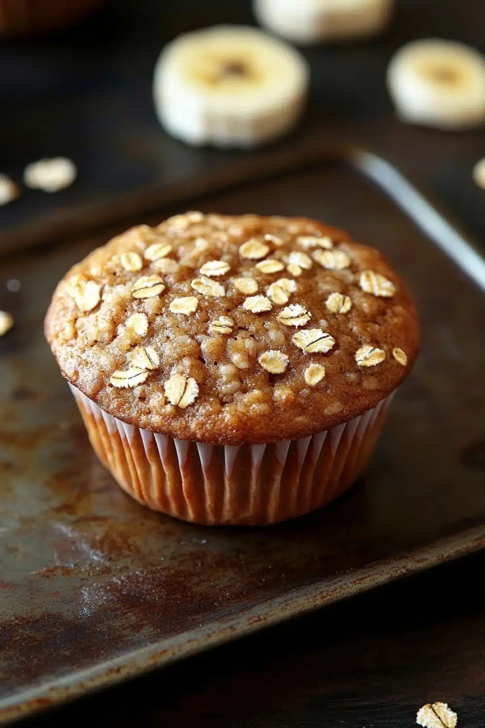 High protein banana oat muffin - the image shows a freshly baked muffin on a baking tray. the muffin is golden brown in color and has a crumbly texture. it is topped with chopped oats, giving it a crunchy appearance. in the background, there are several whole bananas scattered around the tray, adding to the rustic feel of the image.