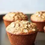 High protein banana oat muffin - the image shows four freshly baked muffins on a baking tray. the muffins are golden brown in color and have a crumbly texture. they are topped with sliced oats, giving them a crunchy appearance. the tray is lined with parchment paper and the background is blurred, making the muffins the focal point of the image.