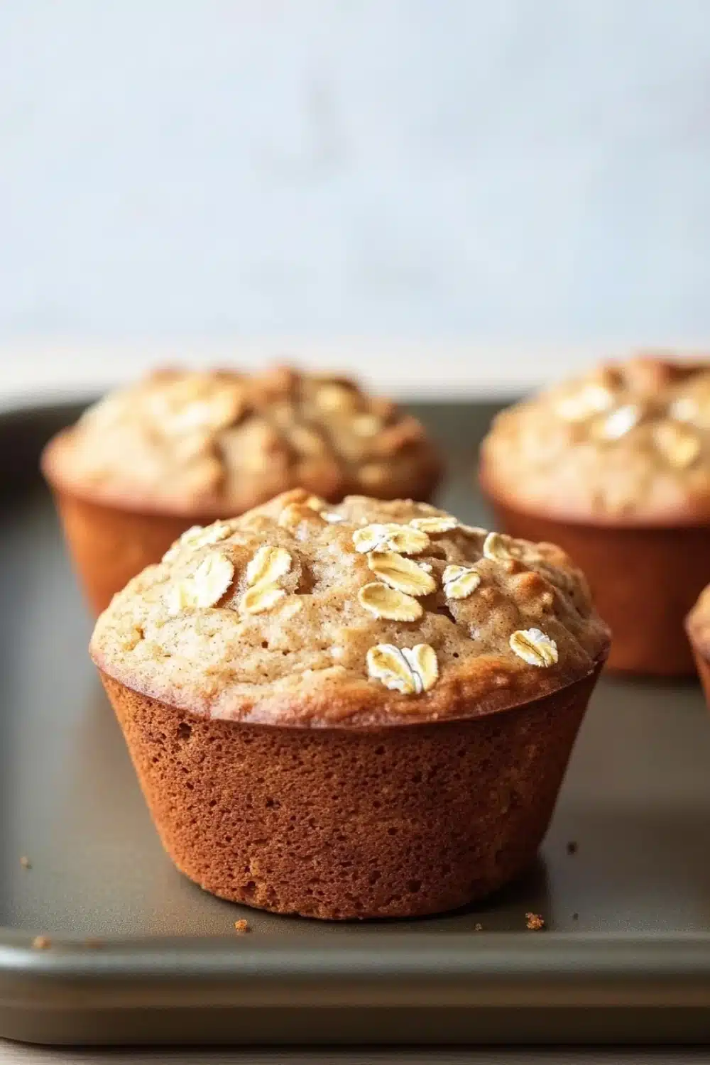 High protein banana oat muffin - the image shows four freshly baked muffins on a baking tray. the muffins are golden brown in color and have a crumbly texture. they are topped with sliced oats, giving them a crunchy appearance. the tray is lined with parchment paper and the background is blurred, making the muffins the focal point of the image.