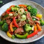High protein beef stir fry - the image is a close-up of a plate of stir-fry with beef and vegetables. the plate is round and has a light brown color with a speckled pattern. the beef is sliced into small pieces and is covered in sesame seeds. the vegetables include broccoli, red bell peppers, yellow bell peppers and green onions. the dish is garnished with sesame seed. the background is a dark grey countertop with small speckles scattered around.