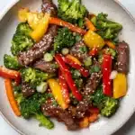 High protein beef stir fry - the image is a close-up of a bowl of stir-fry with beef and vegetables. the bowl is made of light-colored ceramic and is placed on a white marble countertop. the beef is sliced into thin strips and is mixed with various vegetables such as broccoli, red and yellow bell peppers, and green onions. the vegetables are arranged in a colorful and appetizing manner, with some pieces overlapping each other. the dish appears to be freshly cooked and ready to eat.