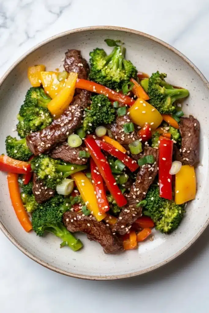 High protein beef stir fry - the image is a close-up of a bowl of stir-fry with beef and vegetables. the bowl is made of light-colored ceramic and is placed on a white marble countertop. the beef is sliced into thin strips and is mixed with various vegetables such as broccoli, red and yellow bell peppers, and green onions. the vegetables are arranged in a colorful and appetizing manner, with some pieces overlapping each other. the dish appears to be freshly cooked and ready to eat.