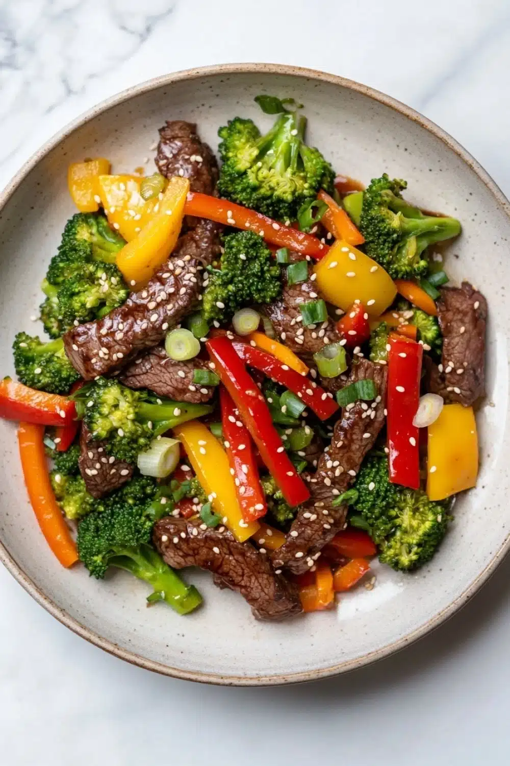 High protein beef stir fry - the image is a close-up of a bowl of stir-fry with beef and vegetables. the bowl is made of light-colored ceramic and is placed on a white marble countertop. the beef is sliced into thin strips and is mixed with various vegetables such as broccoli, red and yellow bell peppers, and green onions. the vegetables are arranged in a colorful and appetizing manner, with some pieces overlapping each other. the dish appears to be freshly cooked and ready to eat.