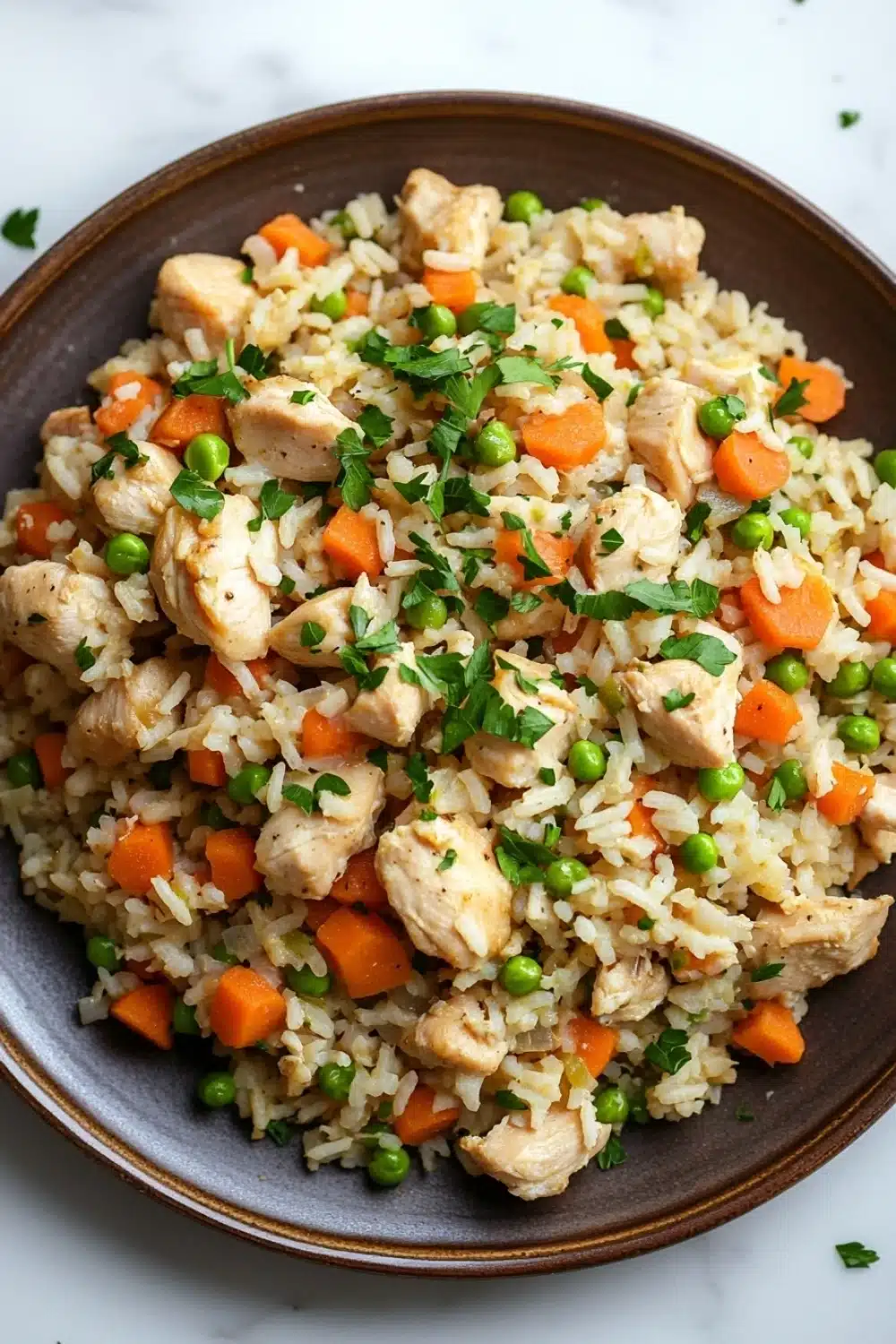 High protein chicken and rice - the image is a close-up of a plate of fried rice with chicken and vegetables. the rice is a light brown color and appears to be seasoned with herbs and spices. the chicken is cut into small pieces and is scattered throughout the dish. there are also small pieces of carrots, peas, and carrots mixed in with the rice. the dish is garnished with a sprig of fresh parsley. the plate is sitting on a white marble countertop.