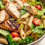 High protein chicken caesar pasta salad - the image is a close-up of a bowl of pasta salad. the bowl is made of ceramic and has a light brown color. the pasta is long and thin, and it appears to be penne. on top of the pasta, there are several pieces of grilled chicken, which are golden brown and appear to be seasoned with herbs and spices. the salad is made up of various greens, including lettuce, cherry tomatoes, and green peas. there is also a sprinkle of parmesan cheese on top. the overall appearance of the salad is fresh and appetizing.