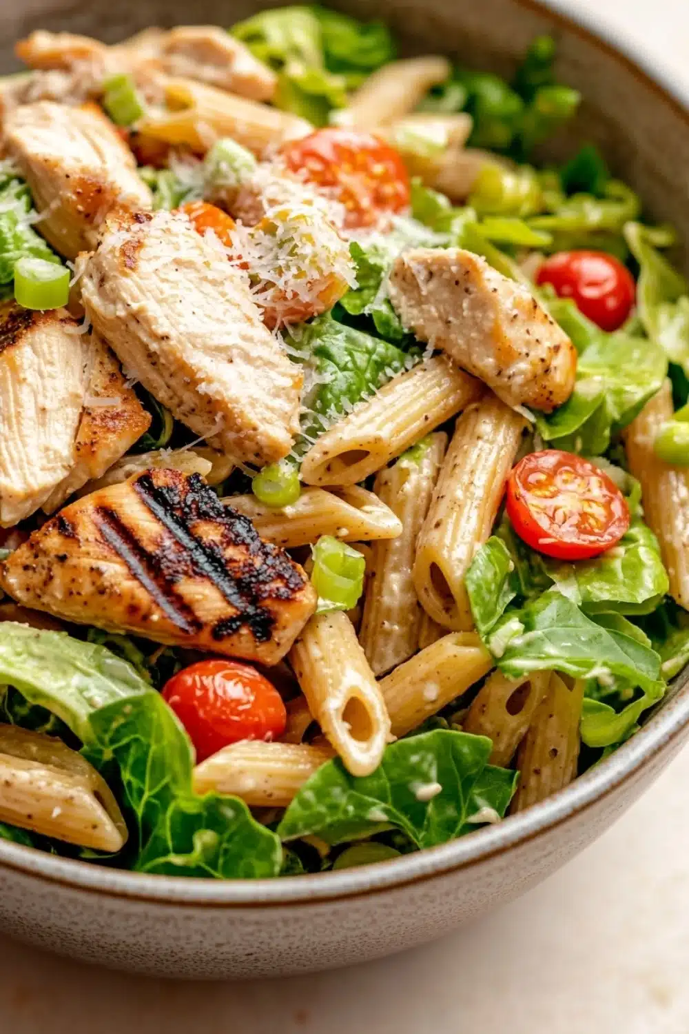 High protein chicken caesar pasta salad - the image is a close-up of a bowl of pasta salad. the bowl is made of ceramic and has a light brown color. the pasta is long and thin, and it appears to be penne. on top of the pasta, there are several pieces of grilled chicken, which are golden brown and appear to be seasoned with herbs and spices. the salad is made up of various greens, including lettuce, cherry tomatoes, and green peas. there is also a sprinkle of parmesan cheese on top. the overall appearance of the salad is fresh and appetizing.