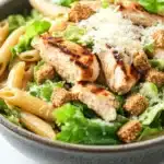 High protein chicken caesar pasta salad - the image is a close-up of a bowl of pasta salad. the bowl is made of dark grey ceramic and is placed on a white marble countertop. the salad is made up of long, thin penne pasta, mixed greens, and crumbled chicken. the chicken is cooked to a golden brown color and appears to be seasoned with herbs and spices. there are also crumbled parmesan cheese scattered throughout the salad, adding a crunchy texture. the overall appearance of the dish is fresh and appetizing.