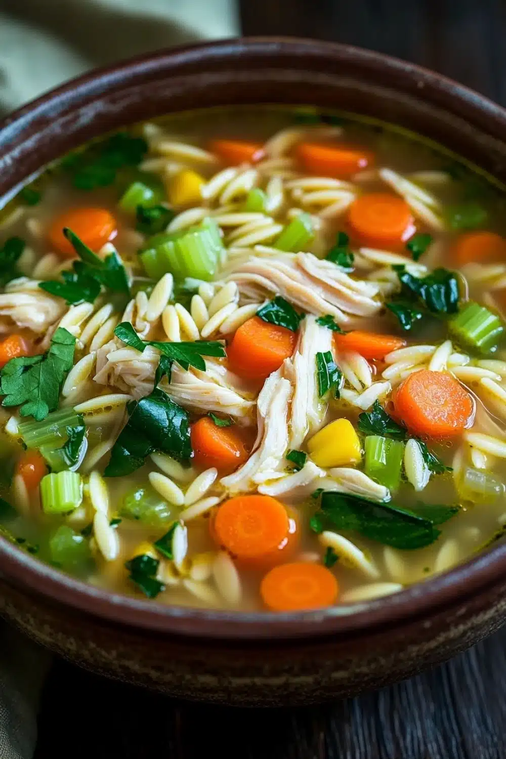 High protein chicken orzo soup - the image is a close-up of a bowl of soup. the soup appears to be a type of noodle soup with chunks of chicken, carrots, celery, and other vegetables mixed in. the bowl is made of ceramic and is sitting on a wooden table. the broth is a light yellow color and the vegetables are a mix of orange, green, and yellow. there are also some sprigs of parsley scattered throughout the soup, adding a pop of green color to the dish. the background is blurred, but it seems to be an indoor setting with a white cloth napkin visible on the side.
