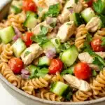 High protein chicken pasta salad - the image is a close-up of a bowl of pasta salad. the bowl is made of light-colored ceramic and is sitting on a white wooden table. the pasta salad is made up of spiral pasta, sliced cucumbers, cherry tomatoes, red onions, and chunks of chicken. the chicken is cooked to a golden brown color and appears to be seasoned with herbs and spices. the salad is garnished with fresh cilantro leaves. the overall appearance of the dish is colorful and appetizing.