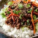 High protein korean beef rice bowl - the image is a close-up of a bowl of food. the bowl is made of ceramic and has a brown rim. the food appears to be a stir-fry dish with a bed of white rice. on top of the rice, there is a mixture of ground beef, sliced carrots, and green onions. the beef is dark brown and looks crispy on the outside. the carrots are bright orange and the onions are bright green. the rice is fluffy and white. the dish is garnished with sesame seeds. the background is dark, making the colors of the food stand out.