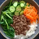 High protein korean beef rice bowl - the image is a close-up of a bowl of food. the bowl is made of a light-colored ceramic material and is placed on a dark grey countertop. on top of the bowl, there is a bed of white rice, which appears to be a type of rice dish. on the side of the rice, there are sliced cucumbers, sliced carrots, and a pile of green spinach leaves. in the center of the dish, there appears to have a generous amount of ground beef or minced meat. the dish is garnished with sesame seeds.