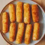 Homemade glutenfree mozzarella sticks - the image shows a plate of nine pieces of breaded fish sticks arranged in a circular pattern on a wooden table. the fish sticks are golden brown in color and appear to be crispy on the outside. they are arranged in two rows of three, with the top row slightly overlapping the bottom row. the plate is white with a light brown rim and has small black speckles around the edges. the background is blurred, but it appears to be a kitchen countertop.