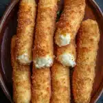 Homemade glutenfree mozzarella sticks - the image shows a brown ceramic plate with six pieces of breaded fish sticks arranged in a circular pattern. the fish sticks are golden brown and appear to be crispy on the outside. they are topped with a dollop of creamy white sauce, which is drizzled over the top. the plate is sitting on a dark grey surface, possibly a table or countertop.