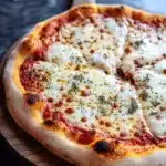 Homemade pizza ingredients - the image is of a freshly baked pizza on a wooden cutting board. the pizza is round and has a golden brown crust. it is topped with melted cheese, red sauce, and various herbs and spices. the cheese is melted and bubbly, and there are small pieces of tomato sauce scattered throughout the pizza. the cutting board is placed on a dark grey countertop, and the background is blurred, making the pizza the focal point of the image.