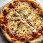 Homemade pizza ingredients - the image is of a freshly baked pizza on a wooden cutting board. the pizza is round and has a golden brown crust. it is topped with melted cheese, red sauce, and small black olives. the cheese is melted and bubbly, and there are small pieces of herbs scattered throughout the pizza. the cutting board is placed on a white marble countertop.