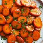 Honey glazed carrots on stove - the image shows a plate of sliced carrots on a white marble countertop. the carrots are bright orange in color and appear to be cooked until they are soft and tender. they are arranged in a circular pattern on the plate, with some overlapping each other. the plate is garnished with chopped parsley, adding a pop of green color to the dish. the background is blurred, making the carrots the focal point of the image.