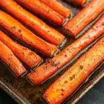 Honey glazed carrots oven roasted - the image shows a tray of cooked carrots on a black surface. the carrots are long and slender, with a golden-brown color and appear to be seasoned with herbs and spices. they are arranged in a single layer on the tray, with some overlapping each other. the tray is lined with parchment paper, and the carrots are glazed with a dark sauce. the background is blurred, but it appears to be a kitchen countertop.