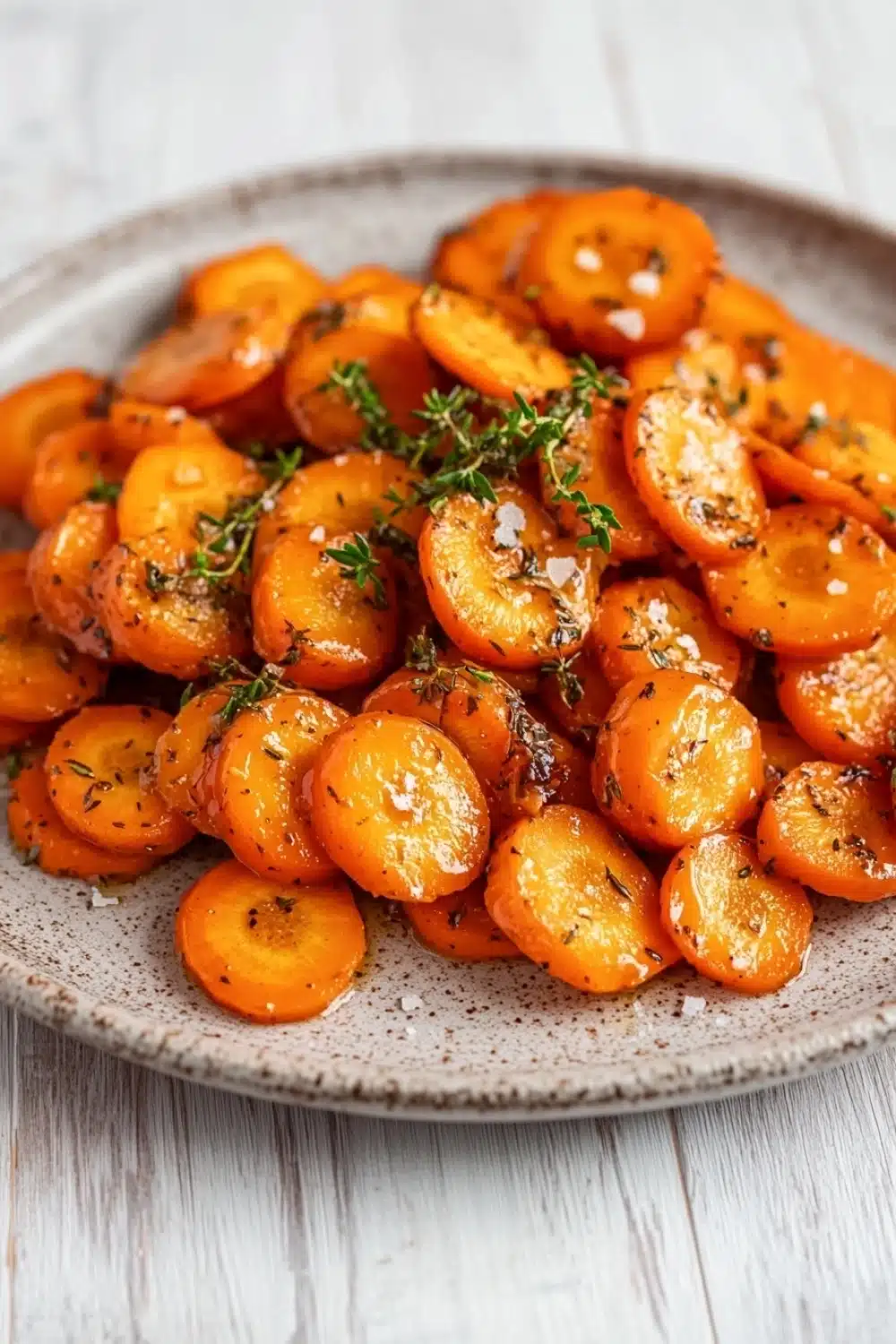 Honey glazed carrots with thyme - the image shows a plate of roasted carrots on a white wooden table. the carrots are orange in color and have a crispy texture. they are arranged in a circular pattern on the plate, with some overlapping each other. there are also some sprigs of fresh herbs scattered throughout the dish, adding a pop of green color to the plate. the plate is made of light-colored wood and has a textured surface. the background is blurred, making the carrots the focal point of the image.
