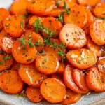 Honey glazed carrots with thyme - the image shows a plate of cooked carrots. the carrots are bright orange in color and appear to be seasoned with herbs and spices. they are arranged in a circular pattern on the plate, with some overlapping each other. the plate is made of light-colored ceramic and has a speckled texture. the background is blurred, but it appears to be a white countertop.
