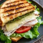 Italian sandwich panini - the image shows a sandwich on a black plate. the sandwich is made with two slices of toasted bread, filled with grilled chicken, cheese, and a generous helping of green pesto sauce. the chicken is cut into thin slices and is arranged in a circular pattern on top of the cheese. the cheese is melted and bubbly, and the pesto is a vibrant green color. the plate is sitting on a wooden table with a blurred background.
