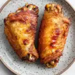Keto chicken wing air fryer - the image shows a plate with two pieces of chicken drumsticks on it. the drumsticks are golden brown and appear to be seasoned with herbs and spices. the plate is round and has a speckled pattern around the edges. the background is a white marble countertop.