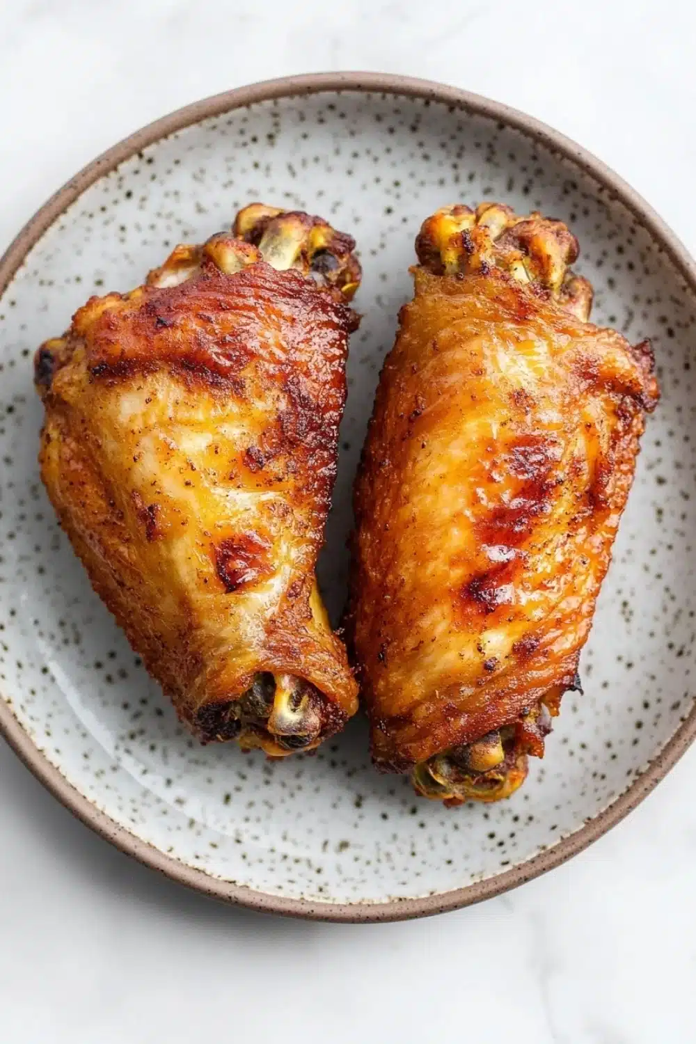Keto chicken wing air fryer - the image shows a plate with two pieces of chicken drumsticks on it. the drumsticks are golden brown and appear to be seasoned with herbs and spices. the plate is round and has a speckled pattern around the edges. the background is a white marble countertop.