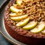 Keto german apple cake - the image is of a round cake on a black plate. the cake appears to be a chocolate tart with a golden brown crust. on top of the cake, there are several sliced pears arranged in a circular pattern. the pears are cut into thin slices and are arranged in an overlapping manner. the top of each pear is sprinkled with chopped nuts, giving the cake a crunchy texture. the plate is sitting on a dark grey countertop.