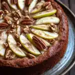Keto german apple cake - the image is of a round cake on a black plate. the cake appears to be a chocolate tart with a golden brown crust. on top of the tart, there are sliced pears arranged in a circular pattern. the pears are cut into thin slices and are arranged in an even layer on top. there are also some pecans scattered throughout the tart. the plate is sitting on a wooden table.